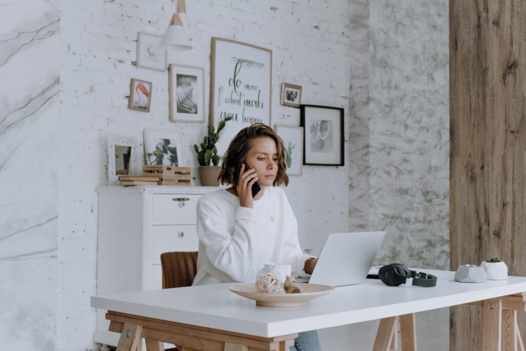 A young woman works from a bright, modern home office, talking on her phone while using a laptop at a white desk in front of a decorated brick wall.