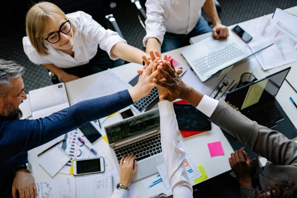 
Gemini said
A diverse team of professionals celebrates a shared achievement by stacking their hands together over a desk cluttered with laptops and project documents.