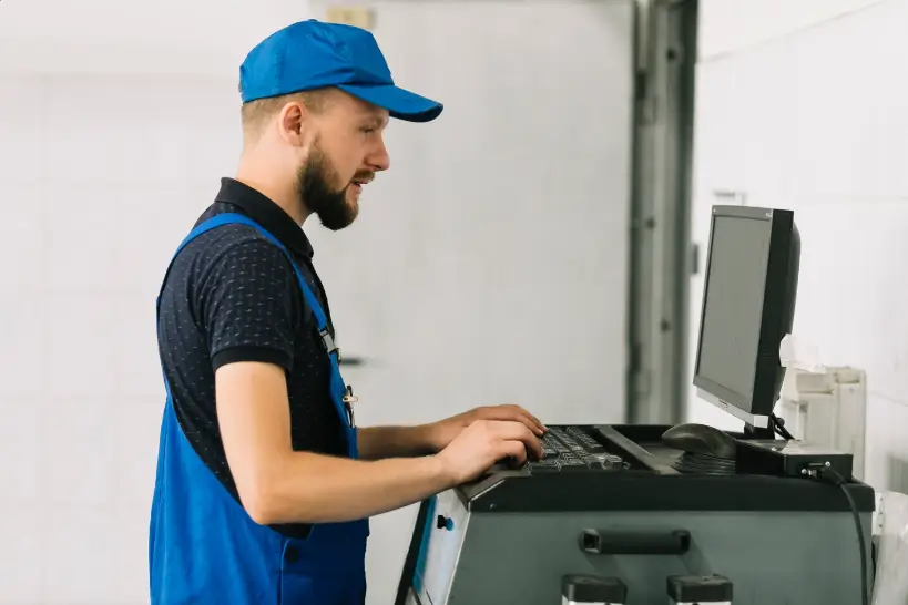 A technician wearing a blue cap and matching overalls operates a computer terminal connected to industrial equipment in a professional workshop environment.